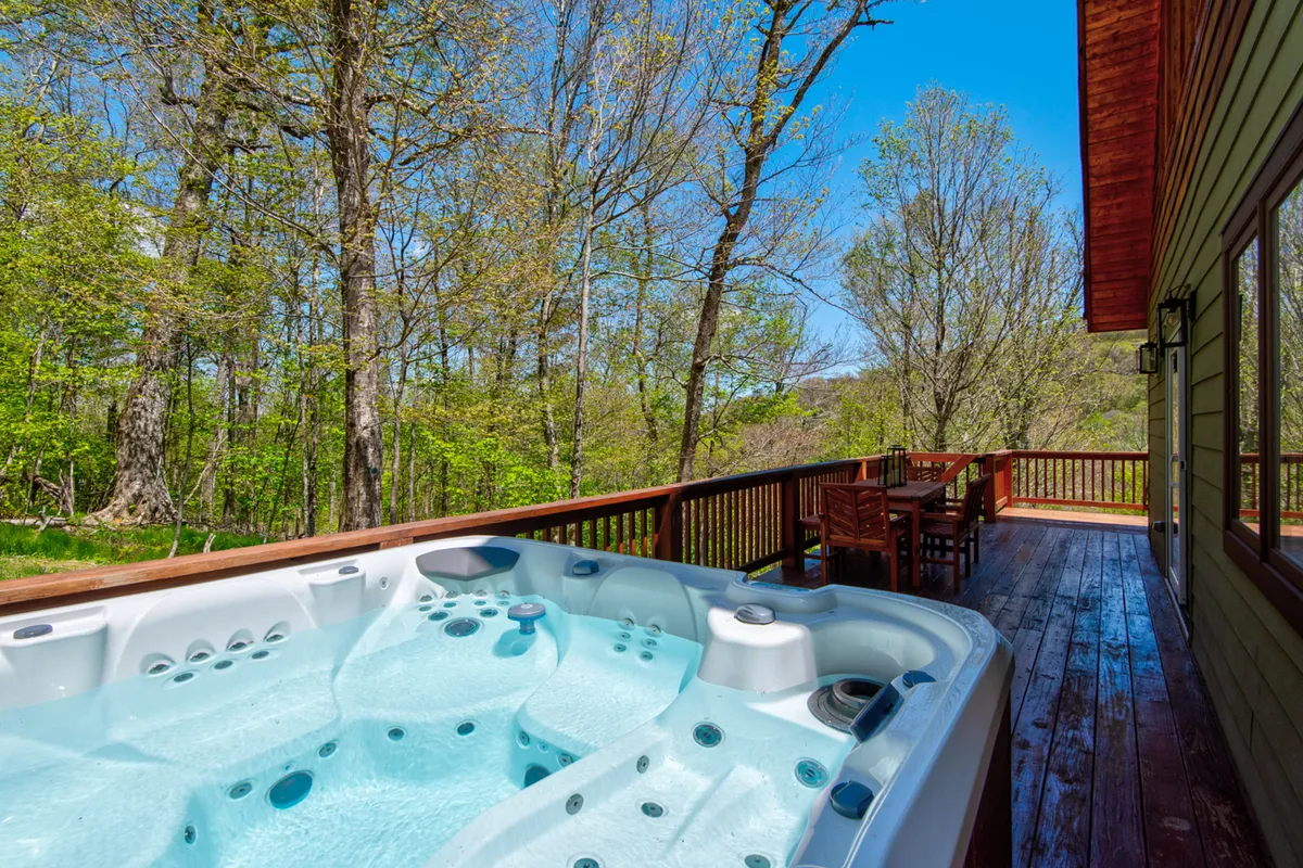 A tranquil outdoor deck with a bubbling hot tub, comfortable dining area, and panoramic views of the surrounding trees, perfectly blending indoor comfort with fresh mountain air.