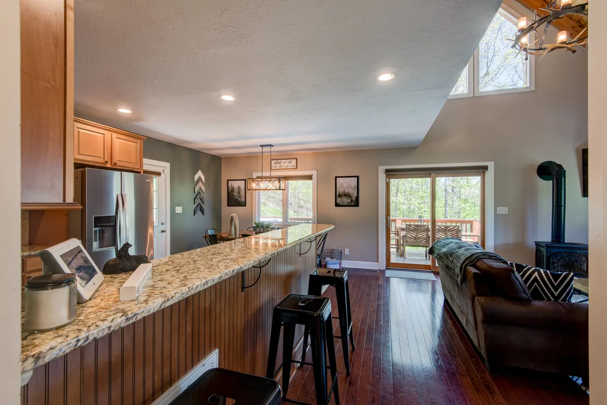 This image provides a view from the kitchen area toward the large windows and a wooden deck, emphasizing both the comfort of the cabin's interior and the inviting outdoor space just outside. The bright natural light and visible deck suggest easy access to open areas perfect for pets and their owners.