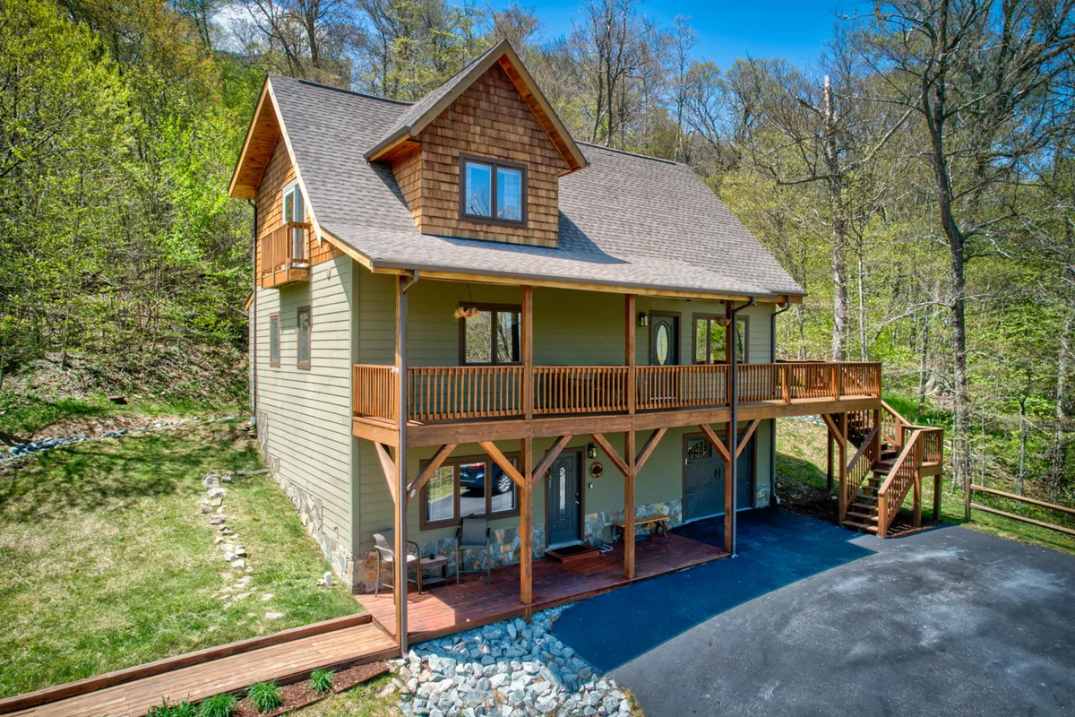 A serene exterior image featuring a wooden balcony with stone foundation, large windows reflecting greenery, and an inviting view of the surrounding mountains and trees. The scene suggests both relaxation and the promise of outdoor adventure.
