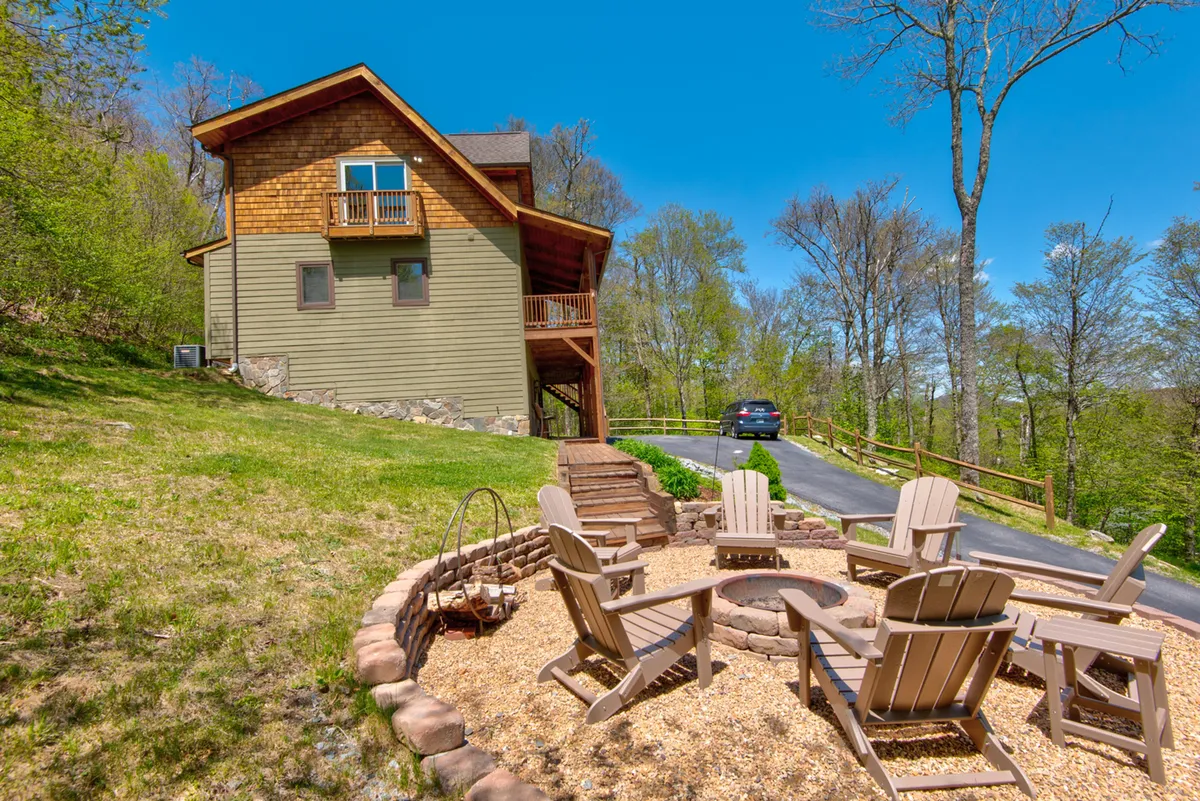 A wide-angle exterior shot showcasing the house, a well-defined stone fire pit surrounded by comfortable seating, and lush trees forming a mountain backdrop. The scene is inviting, illustrating the spaciousness of the yard and the focal point of the fire pit for gatherings under the open sky.