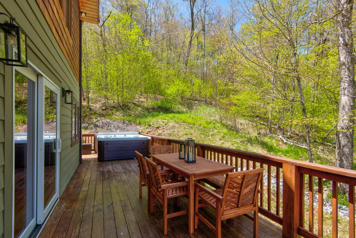 A tranquil exterior deck scene featuring a wooden dining table with chairs, a luxurious hot tub, and lush green surroundings. The deck is spacious, offering both relaxation in the hot tub and outdoor dining with views of the landscape.