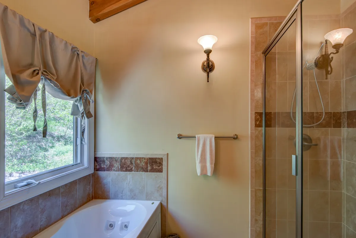 A spa-like bathroom featuring a large soaking bathtub, separate shower stall, window with leafy views, and bright light fixtures. The serene atmosphere invites relaxation and rejuvenation after a long day.