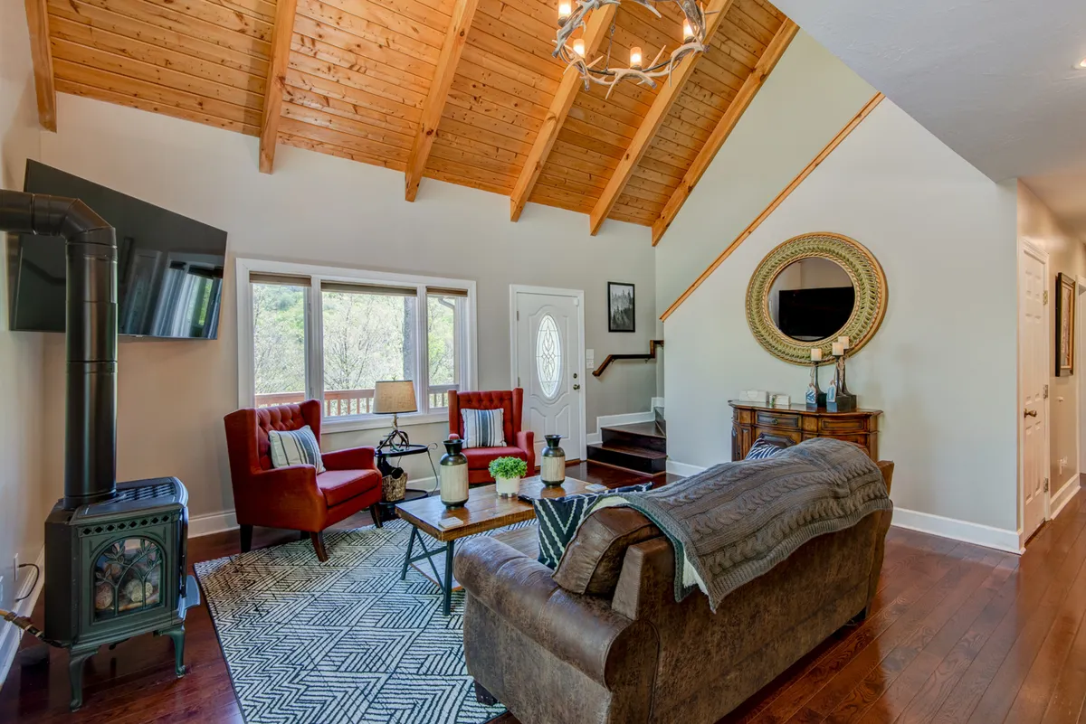 A welcoming main living room with vaulted wooden ceilings, a cozy fireplace, red accent chairs, a central coffee table, and expansive windows that frame the natural mountain scenery outside.