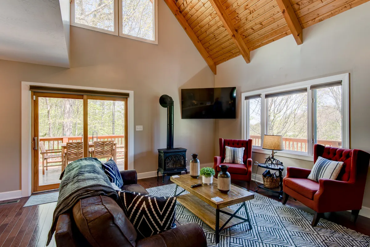 This image showcases a spacious living area with comfortable seating, a high wooden ceiling, and expansive windows that invite abundant natural light. The large windows provide views of the outdoors, hinting at the open spaces and mountain setting surrounding the cabin. The inviting setup suggests a cozy retreat after a day of outdoor adventures with pets.
