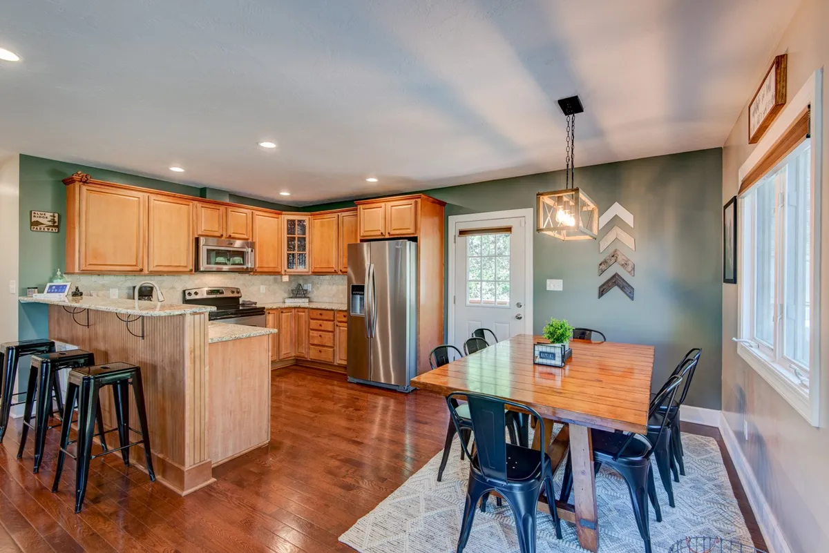 A warm, modern kitchen with a wooden dining table, stainless steel refrigerator, ample cabinetry, and black bar stools—ideal for both culinary creativity and casual breakfasts.