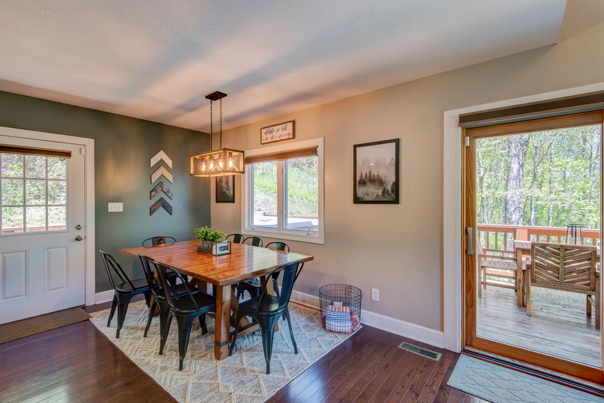 A large wooden dining table sits ready for family feasts or game nights, illuminated by a striking hanging light fixture and surrounded by windows offering serene outdoor views.
