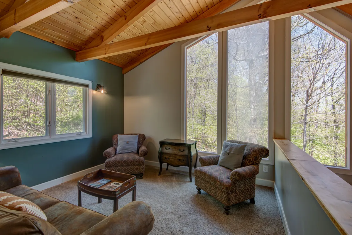 A warm, tranquil living area features comfortable seating, large windows with expansive nature views, and wooden beams overhead. The decorative table and cozy furnishings create an atmosphere ideal for quiet moments or intimate conversation while staying connected to the outdoors.