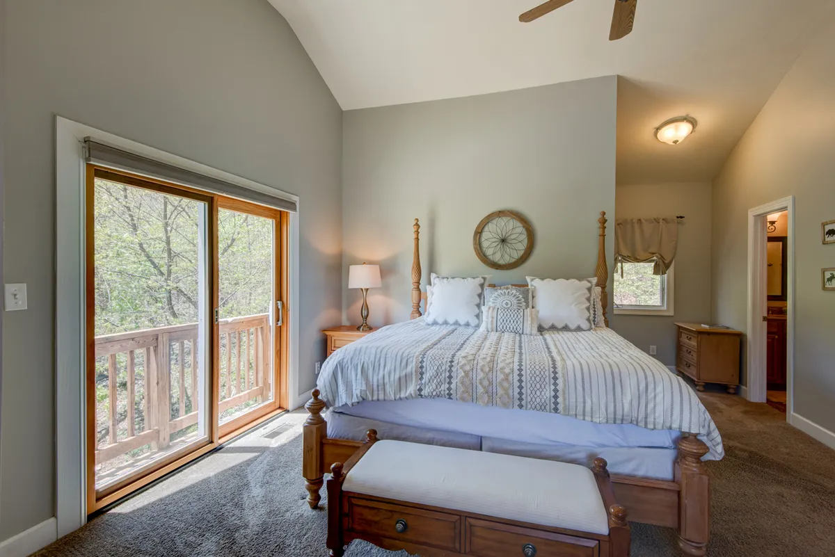A serene bedroom scene shows a large bed positioned to enjoy vistas out a sliding glass door, with a nightstand and decorative elements that add charm. Sunlight spills into the room, inviting guests to wake up and start their day embraced by the mountain landscape.