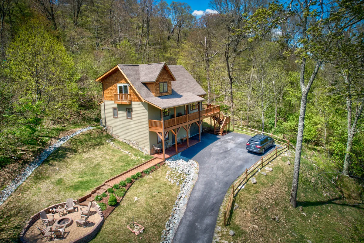 The hero image showcases the exterior of a modern mountain cabin nestled among lush trees, with an inviting fire pit area in the foreground and a spacious driveway leading up to the home. The expansive porch offers the perfect spot to watch a breathtaking sunset, while the fire pit area hints at cozy evenings spent under the stars.